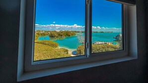 A window with a view of a coastal landscape, including a body of water, greenery, and distant buildings.