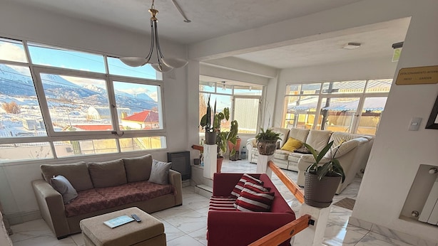 Un amplio salón con un sofá rojo, un sofá beige y un banco de madera. Grandes ventanales ofrecen vistas al exterior.