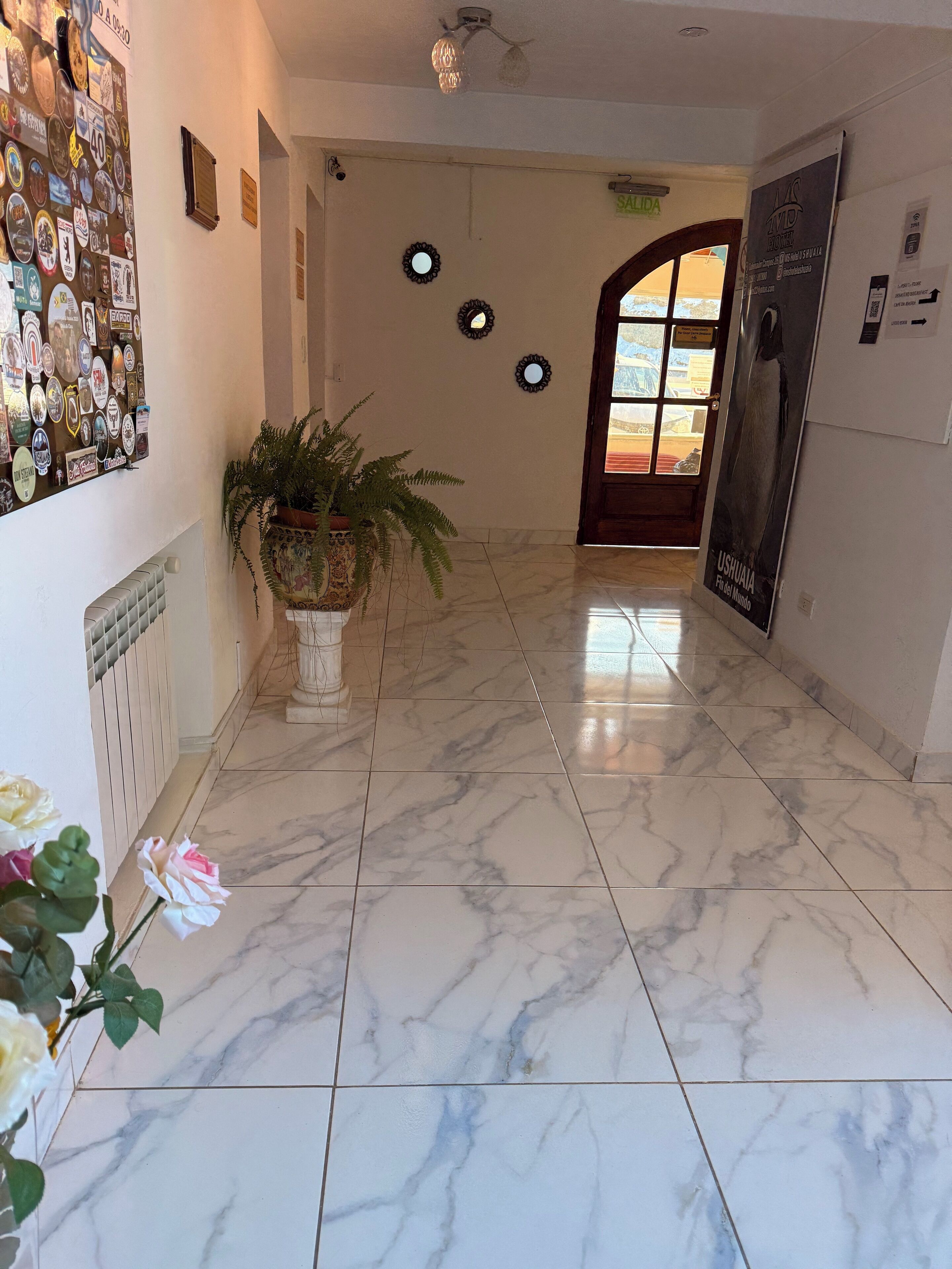 Um hall de hotel com piso de mármore, uma planta em vaso, um painel com diversos adesivos e uma porta com um cartaz.