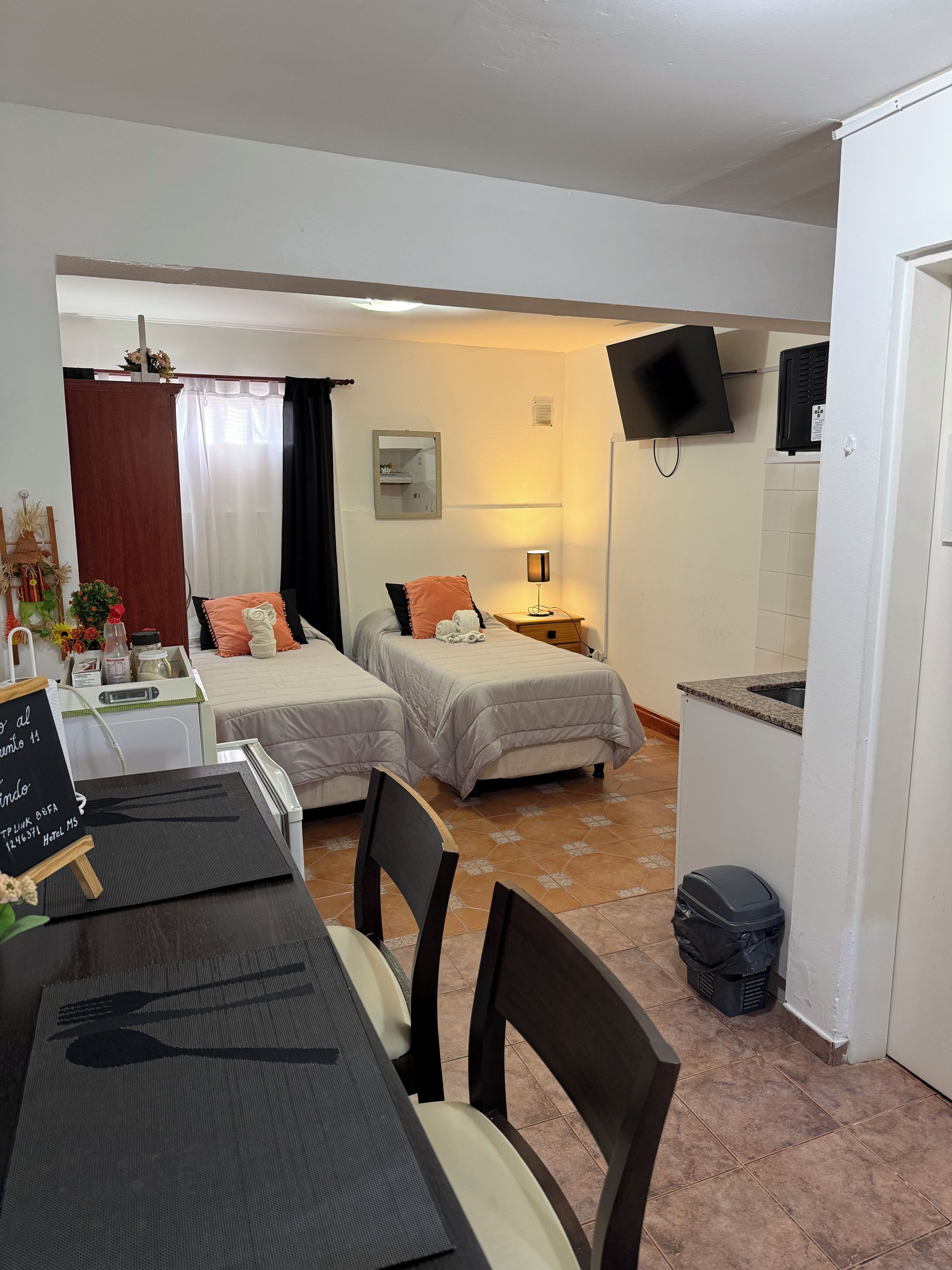 A hotel room with two beds, a dining table, chairs, a TV, and a small kitchen area.