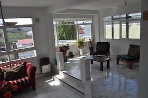 Un salon spacieux avec un canapé rouge, un fauteuil noir et une table basse. De grandes fenêtres offrent une vue sur l’extérieur.
