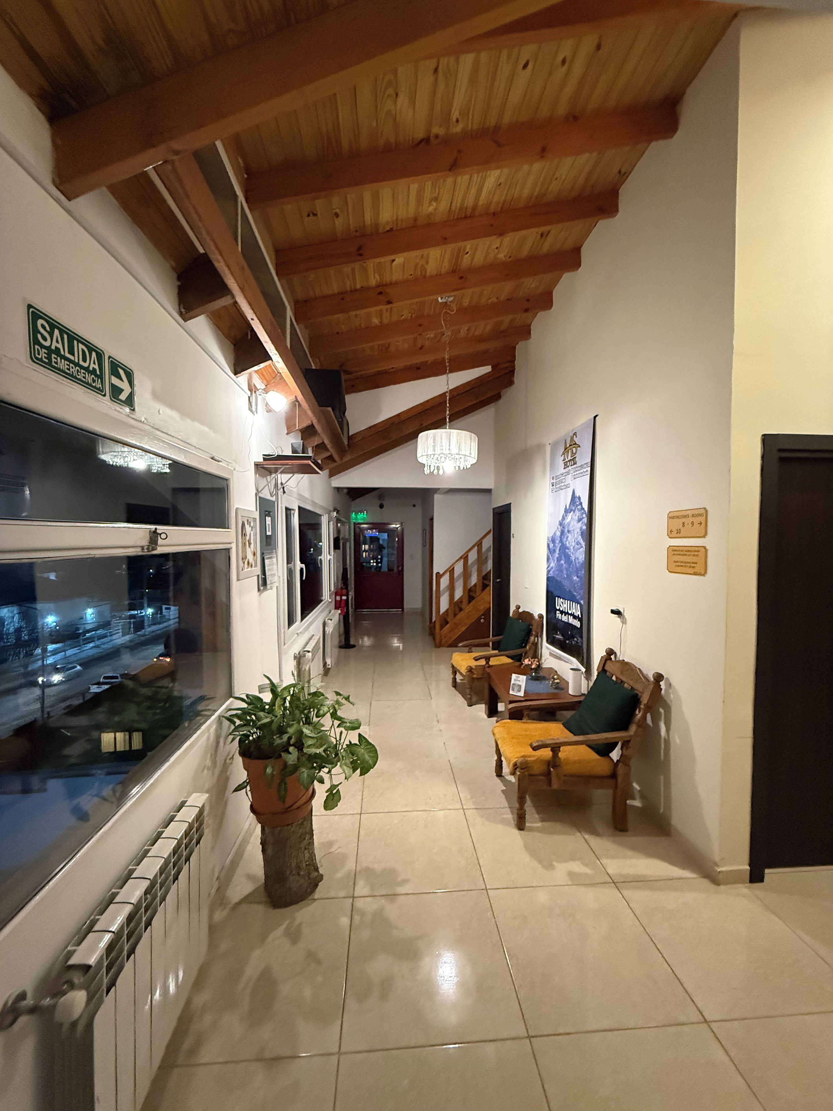 Hall d'albergo con soffitto in legno, reception, area salotto con una panca e sedie, una pianta in vaso e vista sulla strada attraverso una finestra.