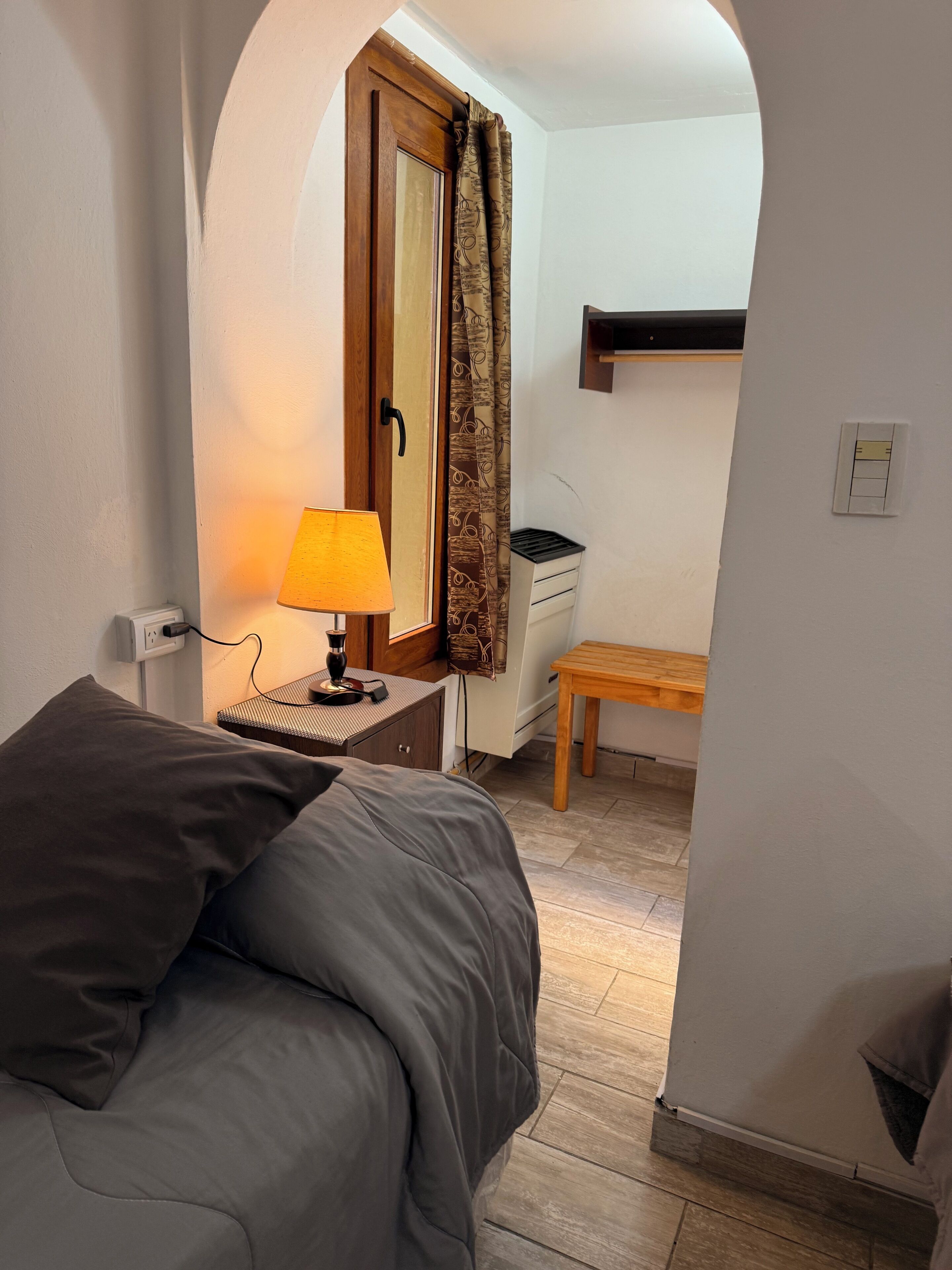A bedroom with a bed, a bedside lamp, a wooden bench, a desk, and a window with curtains.