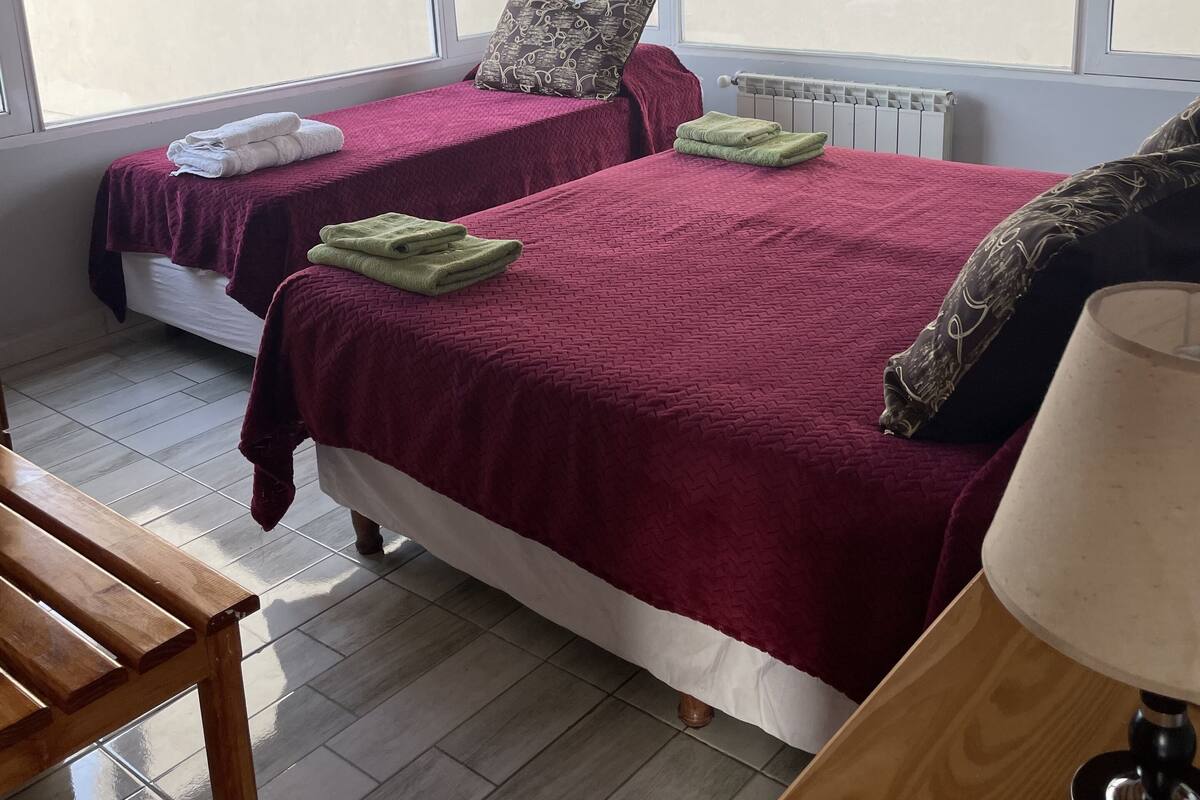 Une chambre avec deux lits, un banc en bois, une lampe et de grandes fenêtres.