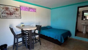 A small room with a bed, a table with two chairs, a wall-mounted shelf, and a window with a decorative item. - Menta Condo (San Felipe)