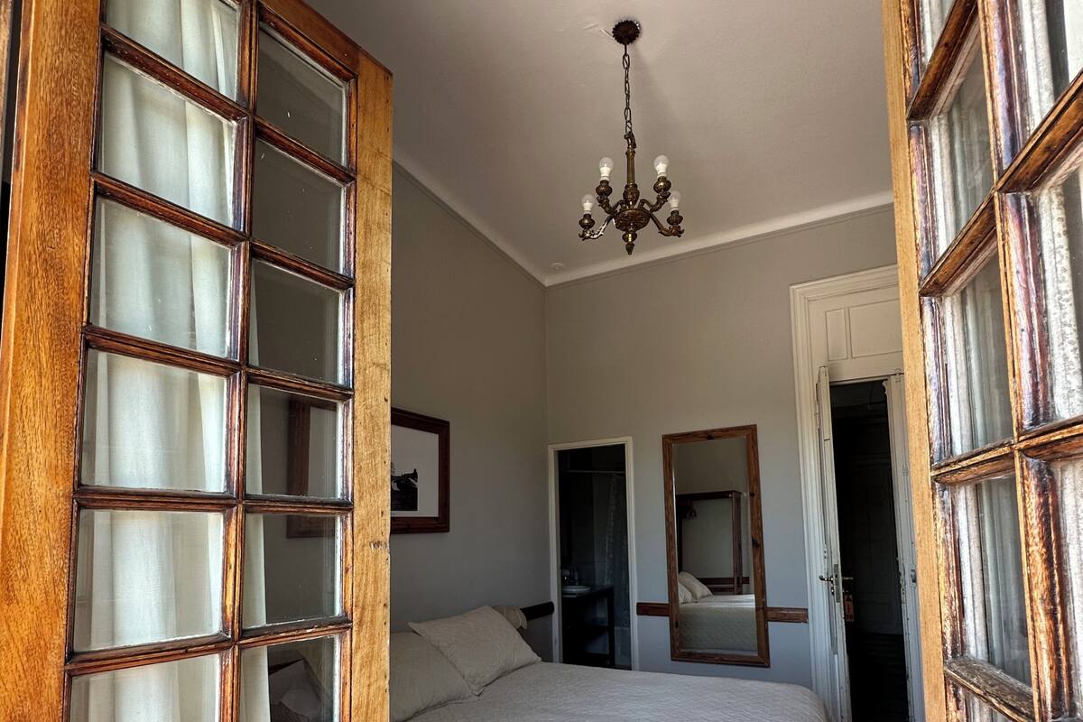 A bedroom with a large bed, wooden-framed windows, a chandelier, and a door leading to another room.