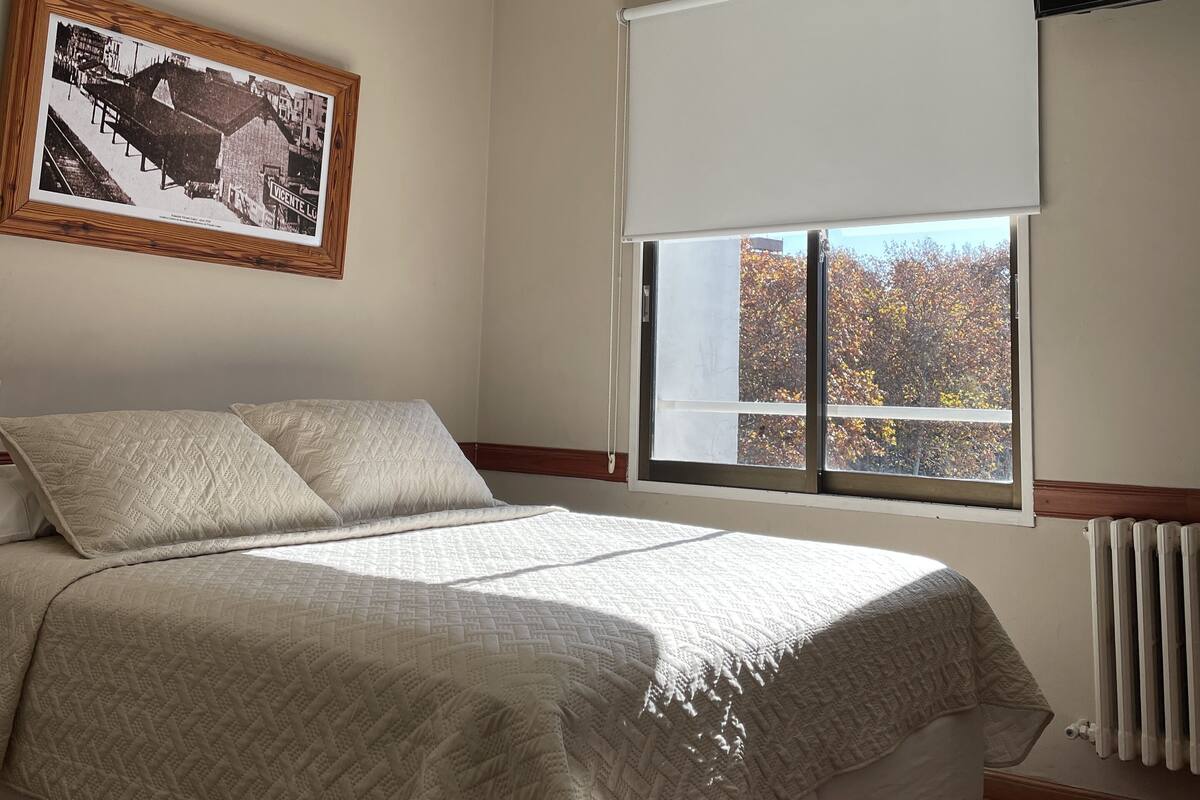 A bedroom with a bed, a window with a view of trees, an air conditioning unit, and a framed picture on the wall.