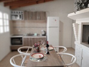 Tavola imbandita con piatti, una bottiglia di vino e un bicchiere. La stanza presenta sedie bianche, un tavolo di legno e una cucina sullo sfondo.