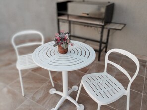 Une petite table ronde, surmontée d’un panier en osier rempli de fleurs, est flanquée de deux chaises blanches à dossier ajouré, le tout posé sur un sol carrelé.