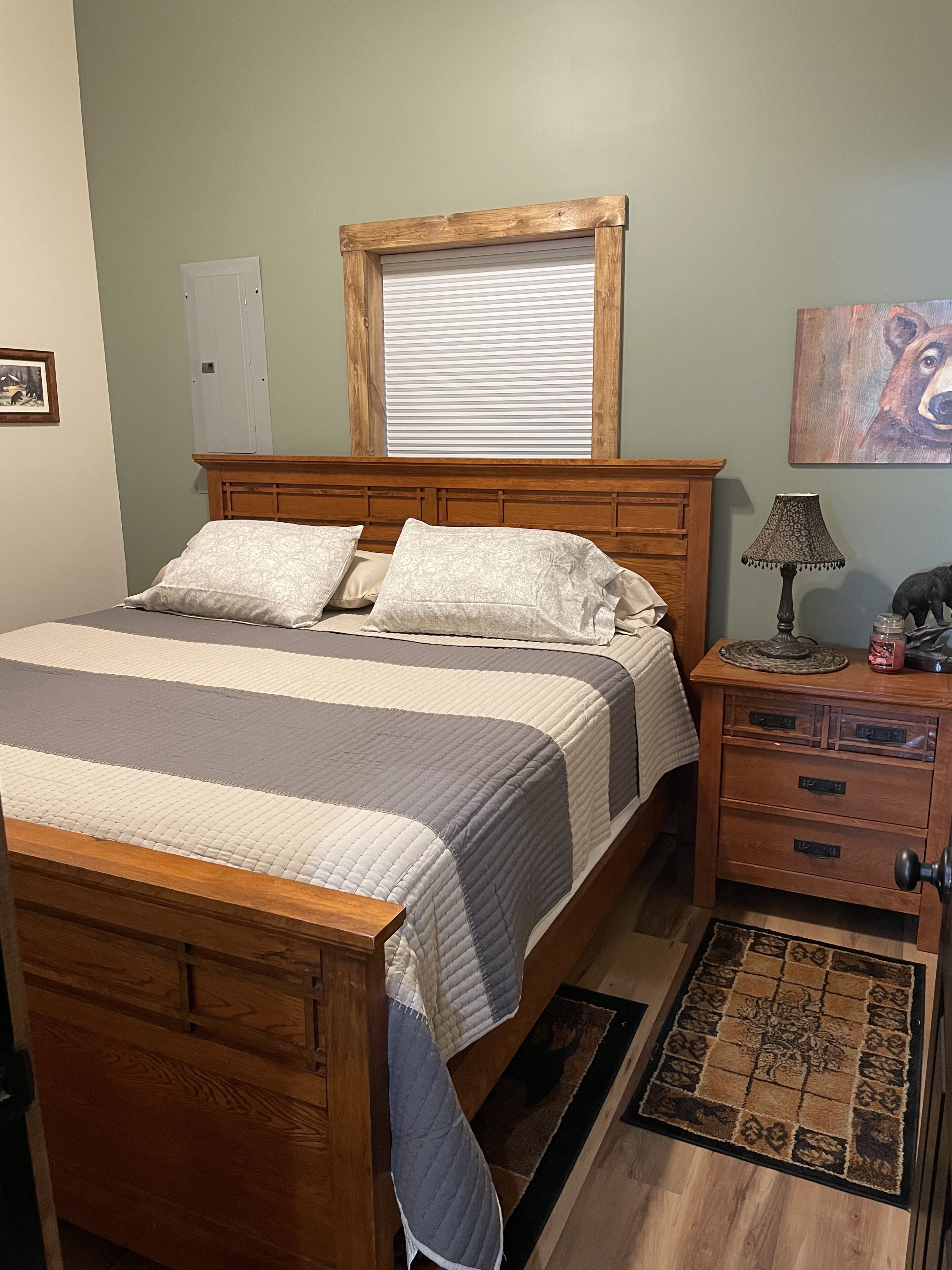 A bedroom with a wooden bed, a nightstand with drawers, a lamp, and a window with blinds.