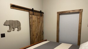 Un dormitorio con una cama, una puerta corredera de madera, un adorno con forma de oso y una ventana con persianas.
