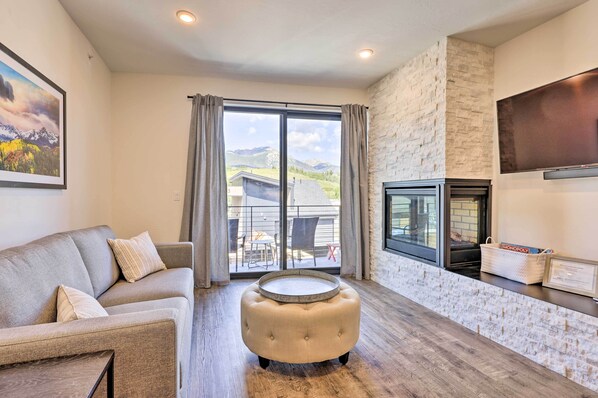 Relax in this serene space with a view of the surrounding mountains, featuring a comfortable sofa, flat-screen TV, and a cozy fireplace.