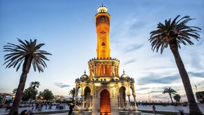 A tall, ornate tower with a clock at the top, surrounded by palm trees and a paved plaza with steps leading up to it. - Maxxmarine hotel (Izmir)