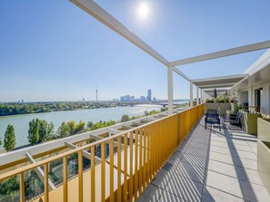 Un balcón con vista al río, mobiliario exterior y un horizonte urbano de fondo.