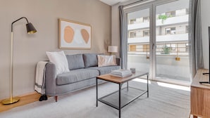 Een moderne woonkamer met een grijze bank, een klein tafeltje, een vloerlamp en een groot raam met uitzicht op een gebouw.