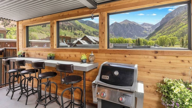 Zona de bar con barra de madera, una parrilla y taburetes, ventanas amplias con vistas a la montaña y una encimera de cocina.