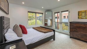 Un dormitorio con cama, cómoda, espejo y balcón con vista a edificios y árboles.