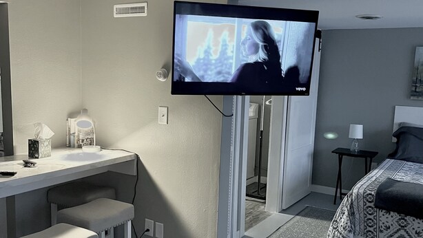A hotel room with a TV mounted on the wall, a desk with two chairs, a bed with patterned bedding, and a small table with a lamp. - Home in the Center of the City! (Alexandria)