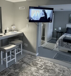 A hotel room with a TV mounted on the wall, a desk with two chairs, a bed with patterned bedding, and a small table with a lamp. - Home in the Center of the City! (Alexandria)