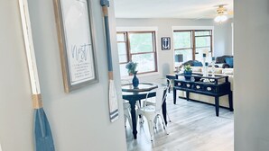 Un salon moderne avec un coin repas, agrémenté d’un panneau décoratif encadré au mur, d’un ventilateur de plafond et offrant une vue sur l’extérieur grâce à de grandes fenêtres.
