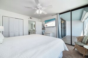 Ein Schlafzimmer mit einem großen Bett, einem Deckenventilator, einer Glastür zu einem Balkon sowie einem Badezimmer mit Spiegel und Waschbecken.
