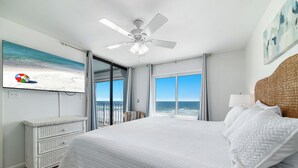 A bedroom with a large bed, a ceiling fan, a beach-themed TV, and a view of the ocean through large windows. - Summerchase 304- Family Friendly Fun Beach Getaway!! (Orange Beach)