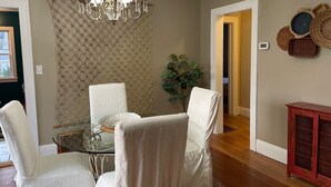 Comedor con mesa de vidrio, sillas blancas tapizadas, lámpara de araña y panel decorativo en la pared.