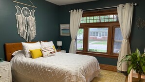 Un dormitorio con cama, mesita de noche, ventana con cortinas, un adorno en la pared y una planta en maceta.