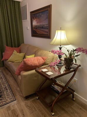 A living room with a beige sofa, patterned cushions, a glass coffee table with a lamp, a painting on the wall, and a green curtain. - Historic Downtown Wilmington . Easy Living by the Water.Apt D  (Wilmington)