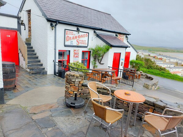 A building with a sign for '7 Licence Diamonds Spirit Stores' and 'Wills Gold Flake Cigarettes'. There are outdoor seating areas with wicker chairs and tables.