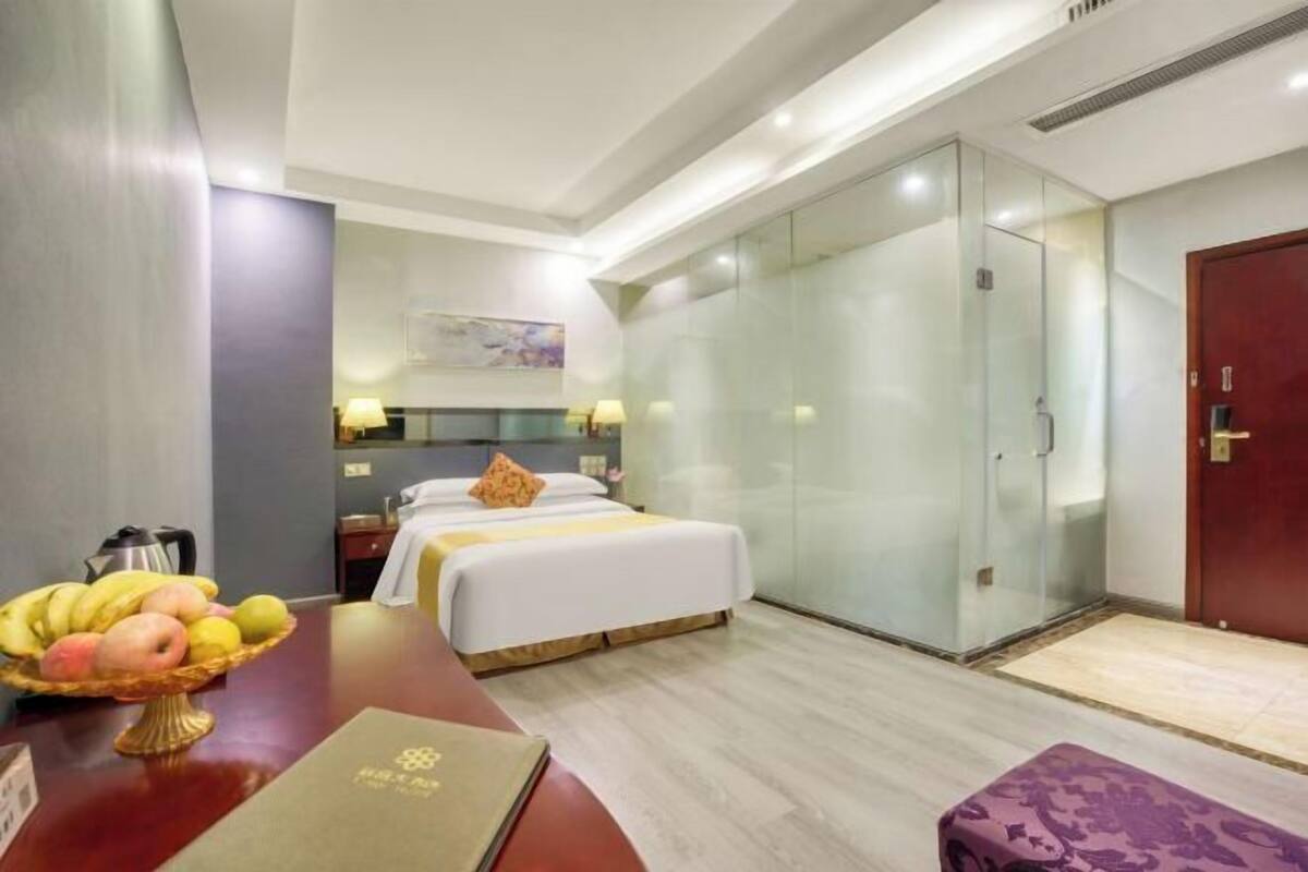 Habitación de hotel con cama, mesita de noche con fruta, lámpara de noche, separador de vidrio, puerta de madera y una alfombra pequeña.