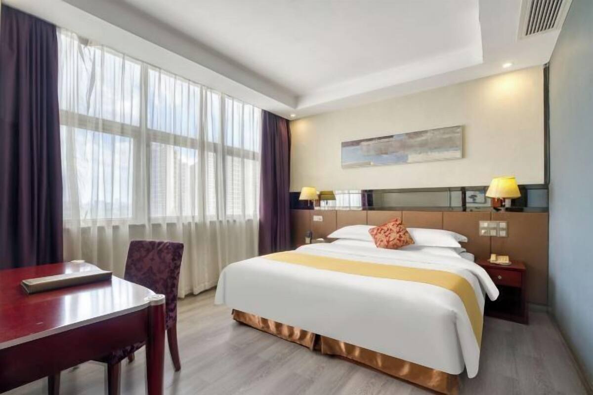 Habitación de hotel con una cama grande, un escritorio, una silla, una mesita de noche y una ventana con cortinas.