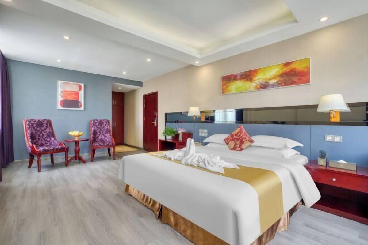 Habitación de hotel con una cama grande, dos sillones, una mesita, una mesita de noche con lámpara, un espejo y obras de arte en las paredes.