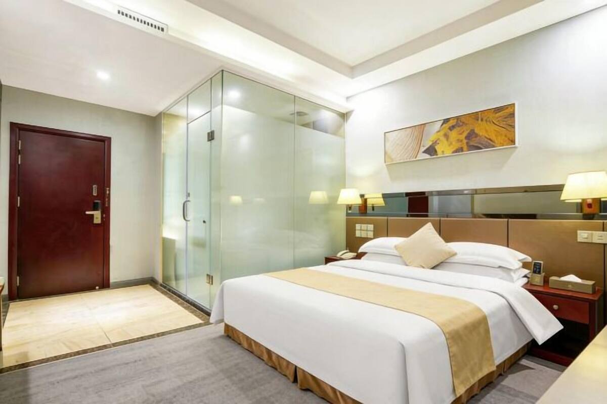 Una habitación de hotel con una cama grande, un armario de vidrio, una puerta de madera y un cuadro en la pared.