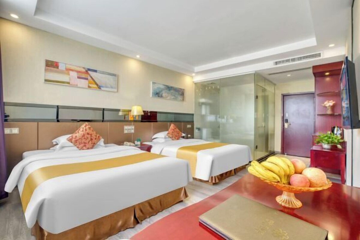 A hotel room with two beds, a glass partition, a desk with a laptop, and a fruit basket.