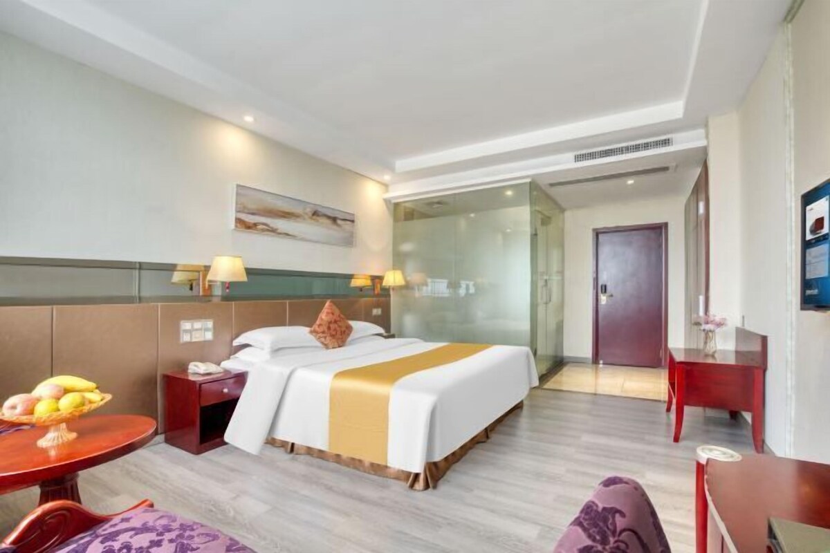 A hotel room with a bed, bedside tables, a desk, a chair, a glass partition, and a door.