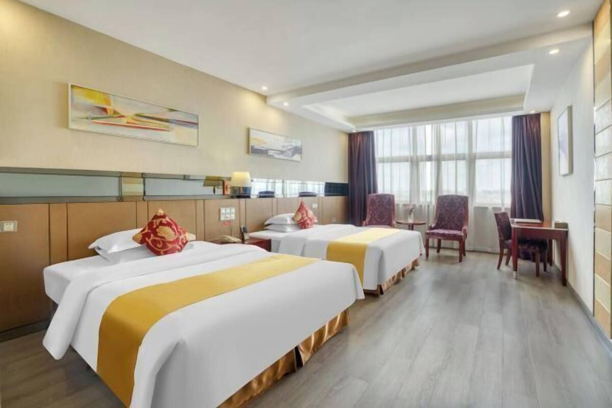Habitación de hotel con dos camas, un escritorio, una silla y ventanas amplias.