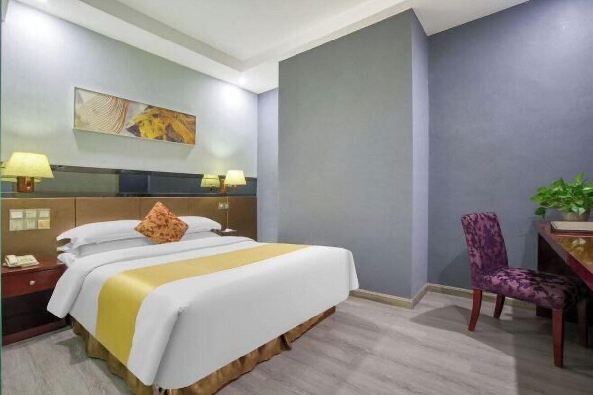 A hotel room with a bed, bedside lamps, a desk with a chair, and a plant.