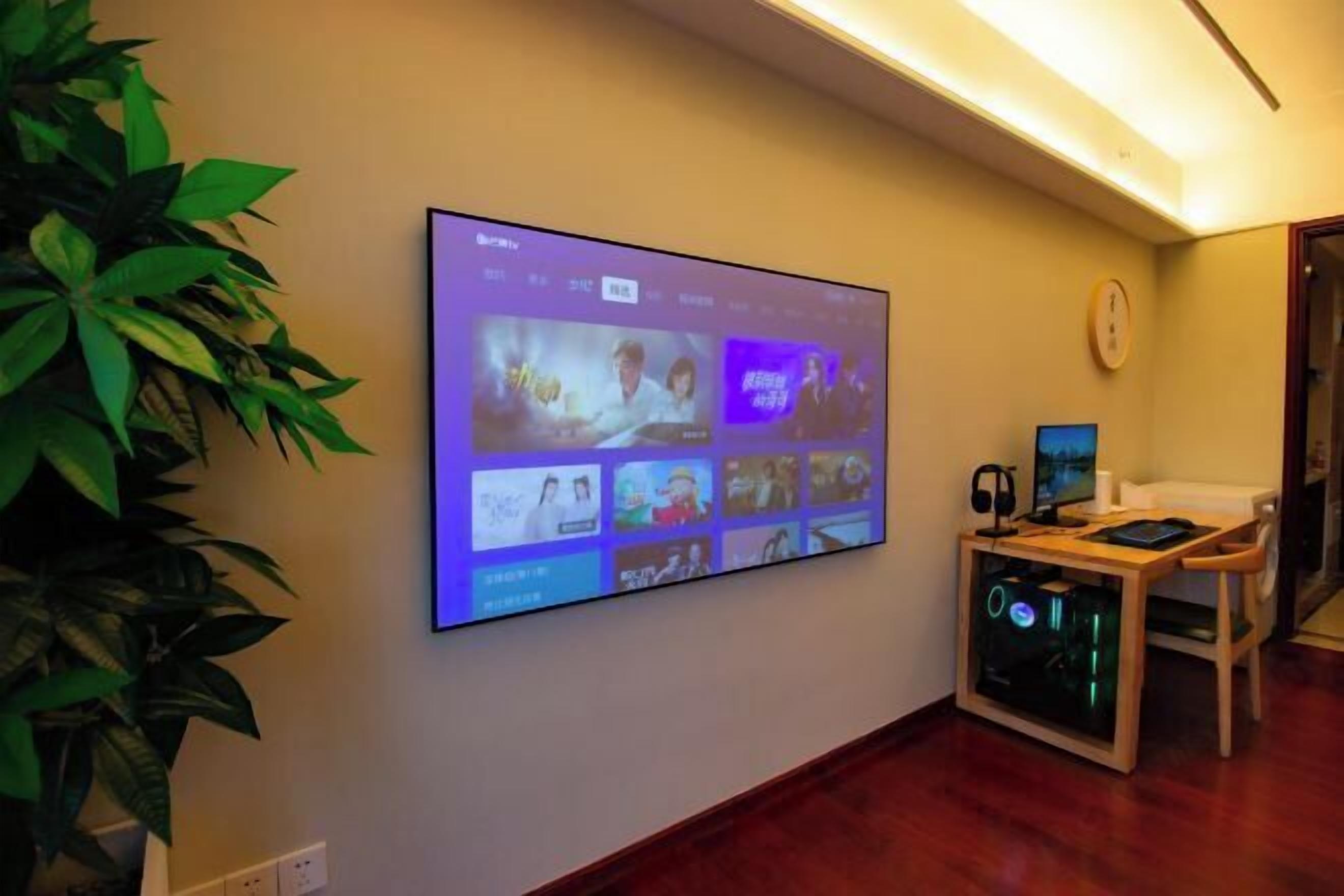 Un écran fixé au mur qui affiche une interface vidéo, un bureau avec un ordinateur, un casque et un écran, ainsi qu’une plante en pot.
