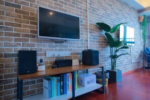 Una habitación con pared de ladrillo, un televisor montado, una estantería de madera con libros y un letrero que dice 'WIFI', además de una planta en maceta.