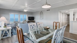 Een moderne woonkamer met een glazen eettafel, witte stoelen, een televisie en uitzicht op zee.