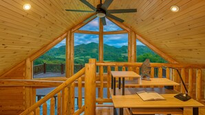 Une cabane en bois avec un plafond en pente, un ventilateur de plafond, une table avec un livre et un portefeuille, et une vue sur des collines verdoyantes par de grandes fenêtres.