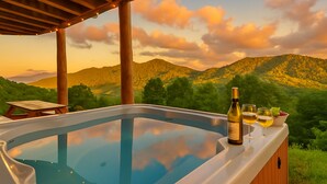 Un bain à remous avec une terrasse en bois, une bouteille de vin et des verres sur une table, le tout dans un décor de montagnes au coucher du soleil.