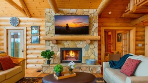 Un intérieur chaleureux de chalet en bois, avec une cheminée en pierre, un téléviseur fixé au mur et des sièges confortables.