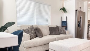 A modern living room with a beige sofa, patterned cushions, a matching ottoman, a white side table, and a large window with blinds. - Newly built guest house with full kitchen and washer/dryer (Simi Valley)