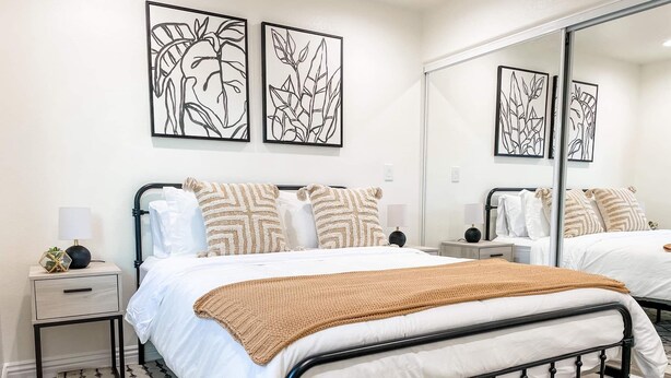 A modern bedroom with a metal bed frame, white bedding, and a patterned rug. There are two bedside tables with lamps, a large mirror, and wall art above the bed. - Newly built guest house with full kitchen and washer/dryer (Simi Valley)