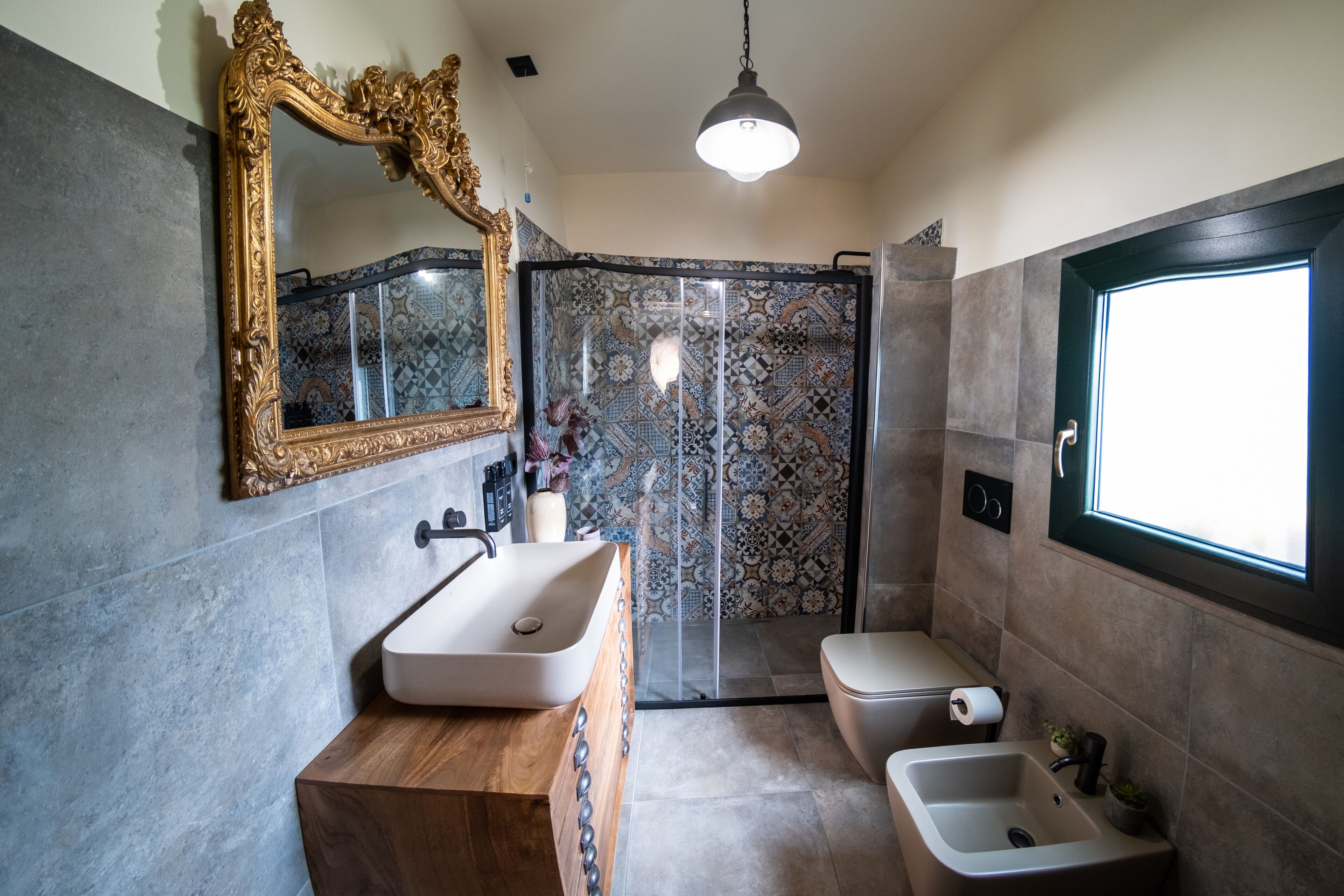 A bathroom with a wooden vanity, a large mirror, a shower with patterned tiles, and a window.