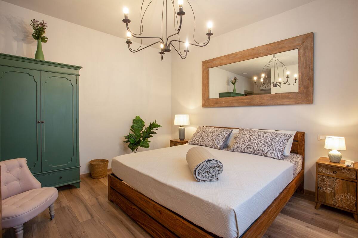 A bedroom with a wooden bed, a green wardrobe, a chair, a mirror, and a chandelier.