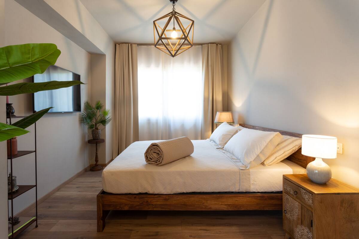 A neatly made bed with a rolled towel, a bedside lamp, and a geometric hanging light fixture in a room with a wooden floor and a large plant.