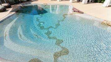Una piscina trasparente con fondo di ciottoli, circondata da lettini e un pergolato.